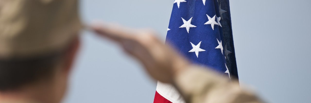 View of the back of a US Army Soldier in uniform, saluting to the american flag. the soldier is blurred, and the flag is in focus