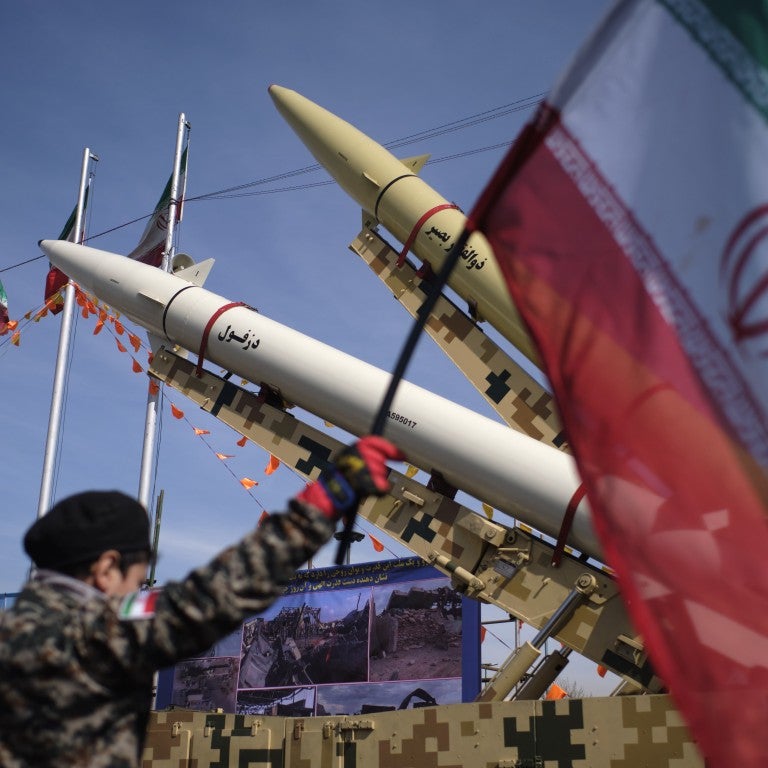 Young Iranian boy wearing Islamic Revolutionary Guard Corps (IRGC) uniform holds Iran flag in front of missile 