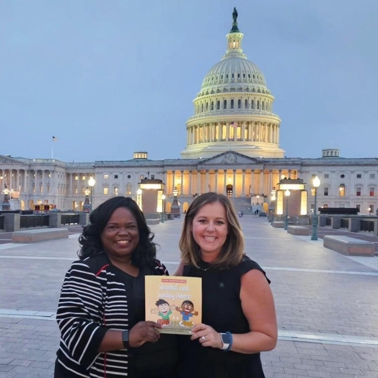 Left - Candace Bazemore. Right - Gabby Leon Spatt, holding the book "Shabbat and Sunday Dinner". Both are smiling in front of the Capitol building in Washington, DC.
