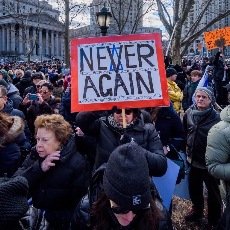 Photo of a crowd and a woman holding a sign saying "Never Again"