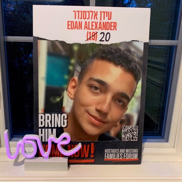 Edan Alexander hostage missing persons poster. red text reading Edan Alexander in Hebrew and English, 19 crossed out and 20 is drawn on. Below is a photo of a smiling young man. The poster is leaning on a windowsill and a small purple "love" sculpture is placed in front of the poster.
