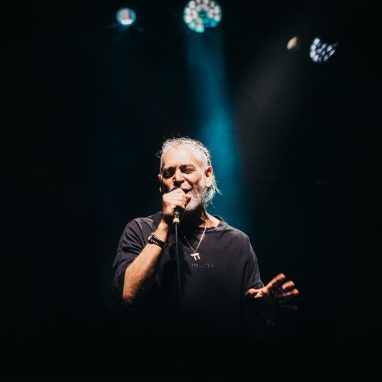 Singer Matisyahu on stage singing into a microphone, background is dark except for one spotlight directed down towards him. Matisuahu is wearing a dark shirt and a large silver necklace with a Chai pendant.