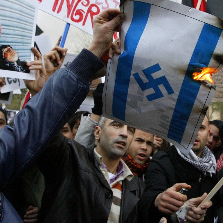 Photo of a man burning a flag that resembles the Israeli flag, but with a Swastika rather than a Star of David